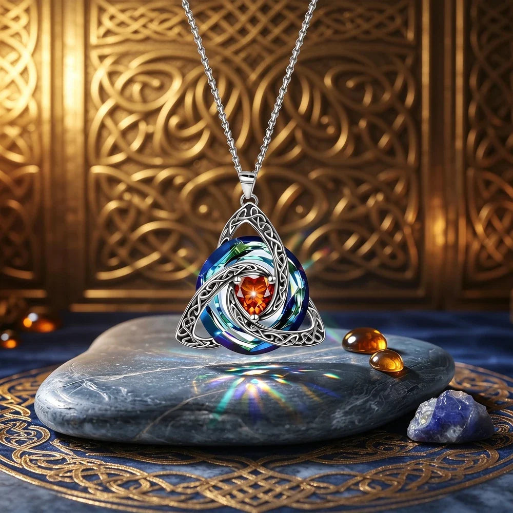 A silver Celtic knotwork pendant necklace with a swirling blue and white iridescent inlay and a central orange heart-shaped gemstone, displayed on a dark stone slab with decorative props against a gold patterned background.