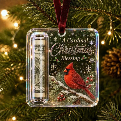 A clear acrylic Christmas ornament hanging on a Christmas tree, featuring a red cardinal, snowflakes, the text 'A Cardinal Christmas Blessing,' a rolled dollar bill in a tube, and a dark red hanging ribbon.