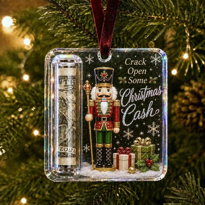 A Christmas ornament hanging on a tree branch, featuring a nutcracker figure, a rolled dollar bill inside a clear tube, and the text 'Crack Open Some Christmas Cash' with gift boxes and snowflakes on a dark background.