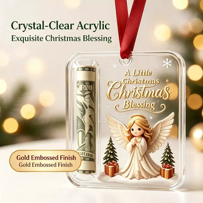 A clear acrylic Christmas ornament with a rolled dollar bill compartment and an embossed angel design with the text 'A Little Christmas Christmas Blessing.' The ornament hangs from a red ribbon against a softly blurred holiday background with warm bokeh lights.