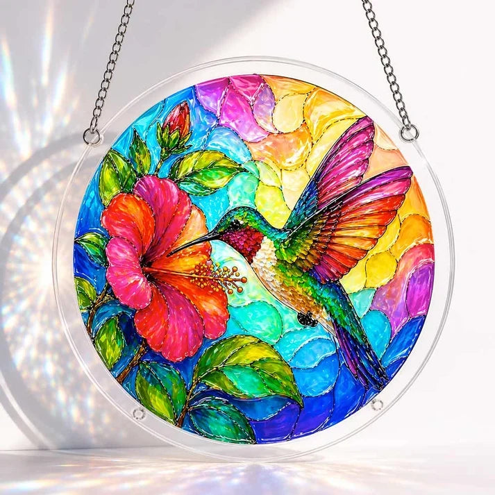 A round acrylic suncatcher with a colorful hummingbird and hibiscus flower in a clear ring with metal chains laid on a flat surface