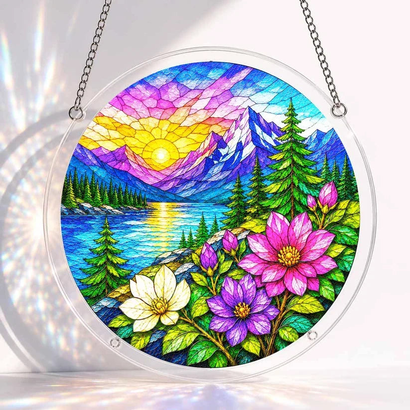 A round acrylic suncatcher with a stained glass style alpine sunrise lake mountain and pink purple flowers hanging on a chain