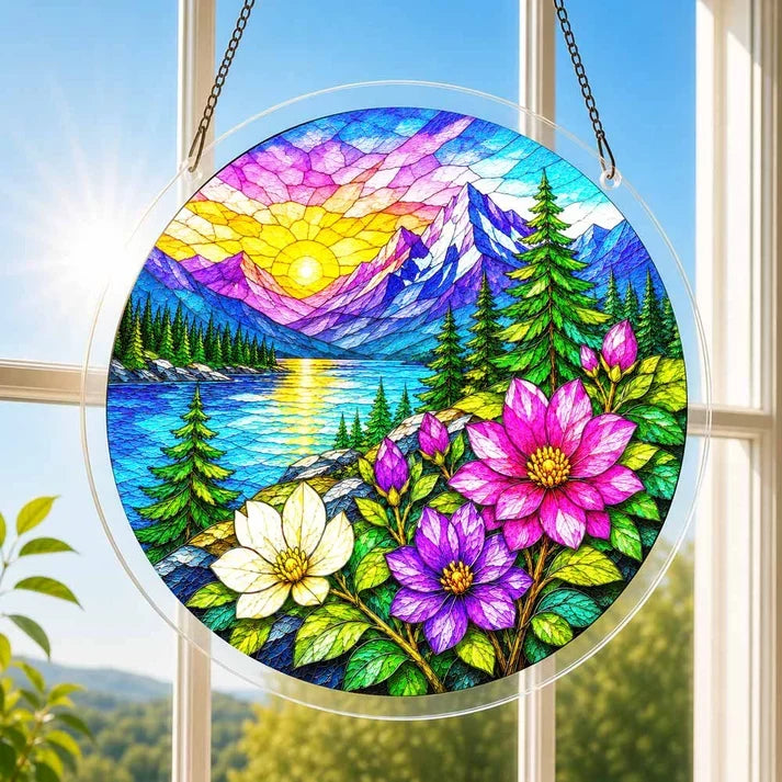 A round acrylic suncatcher with an alpine sunrise snowy peaks a lake pine trees and flowers hanging from a chain in front of a bright white window