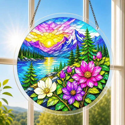 A round acrylic suncatcher with an alpine sunrise snowy peaks a lake pine trees and flowers hanging from a chain in front of a bright white window