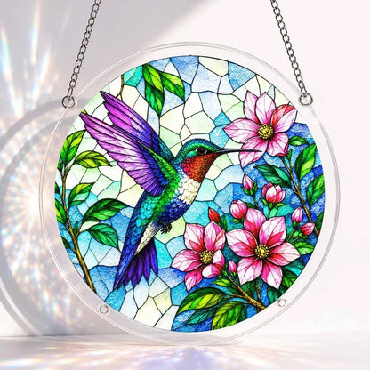 A round acrylic suncatcher featuring a colorful hummingbird beside pink blossoms on a blue stained glass background