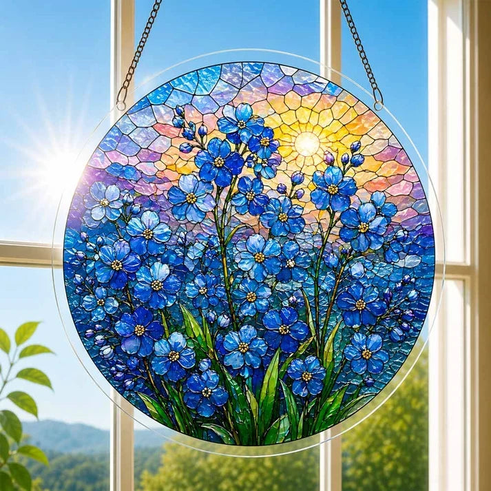 A round acrylic suncatcher with blue forget me not flowers and a glowing sun hanging from a chain in front of a bright white window