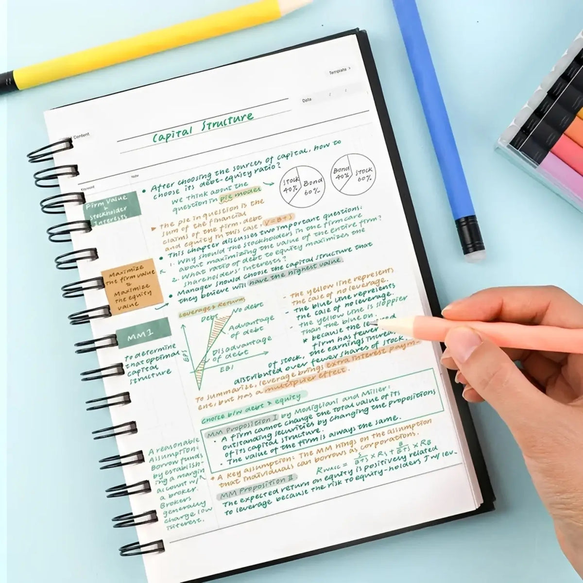 A hand writes with a coral gel pen on a colorful highlighted spiral notebook placed on a light blue desk