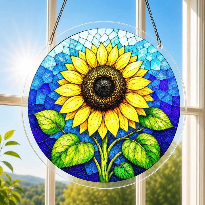 A round acrylic suncatcher with a large golden sunflower and green leaves hanging from a chain in front of a bright white window