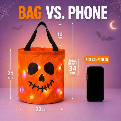 An orange pumpkin-shaped Halloween bucket bag with black handles and a jack-o'-lantern face, shown with size measurements and a phone for a size comparison on a purple Halloween-themed background.