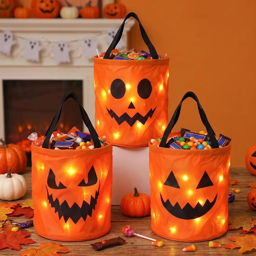 Three orange jack-o'-lantern-shaped fabric candy baskets with black handles and built-in warm white string lights, filled with assorted candies, displayed on a table in a Halloween-themed room with pumpkins and autumn leaves.