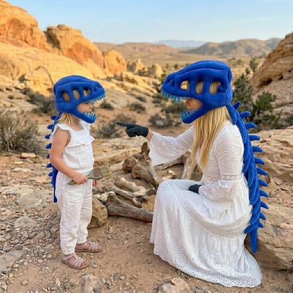 A woman and a child wearing matching blue dinosaur costume headsets and spiky back accessories pose outdoors in a rocky desert landscape. The woman wears a long white dress and black gloves, while the child wears a white sleeveless outfit.
