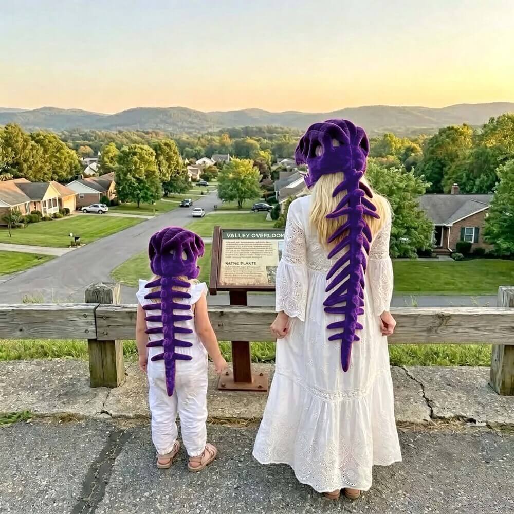 A woman and a child wearing matching purple dinosaur skeleton costumes, viewed from behind as they stand outdoors at a scenic overlook with a town and mountains in the distance.