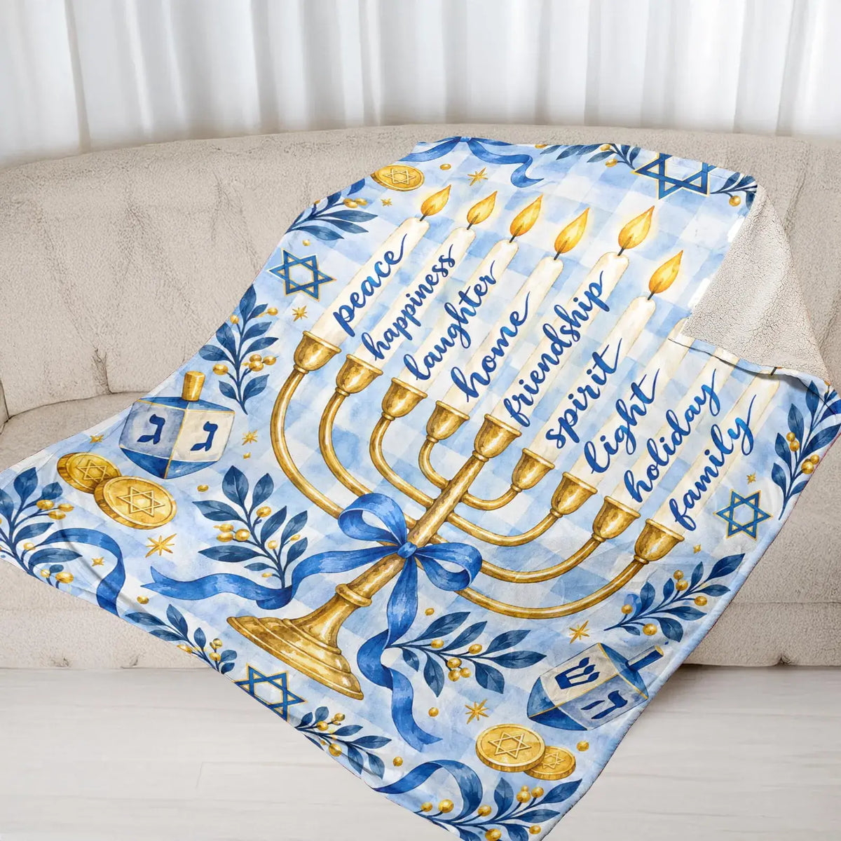 The watercolor Hanukkah blanket featuring a golden menorah and dreidels is draped over a beige sofa with striped curtains behind