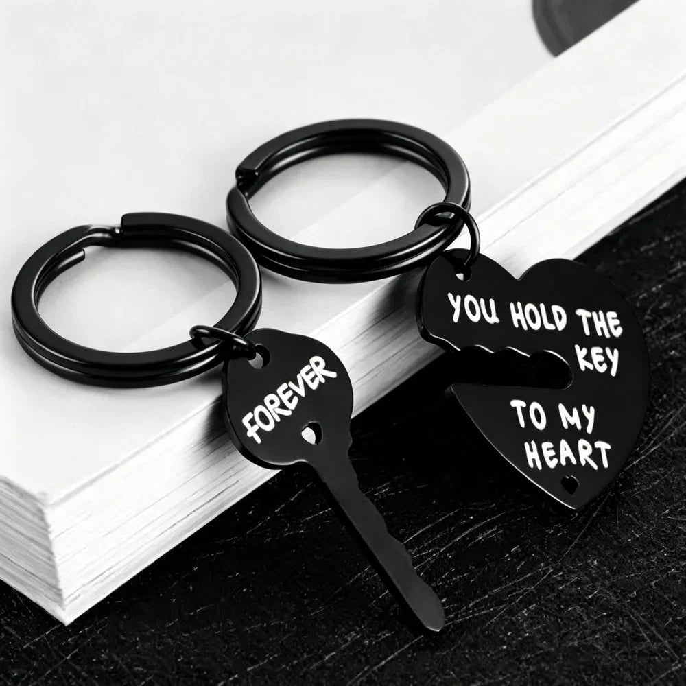 Two black metal keychains shaped like a broken heart and key, with white engraved text reading 'FOREVER' and 'YOU HOLD THE KEY TO MY HEART', displayed on stacked white books over a dark surface.