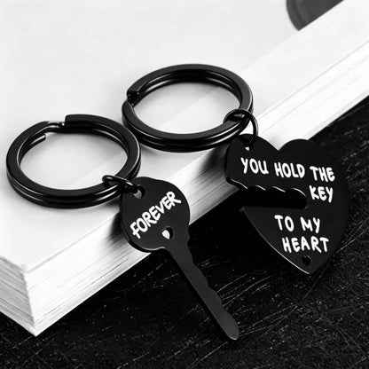 Two black metal keychains shaped like a broken heart and key, with white engraved text reading 'FOREVER' and 'YOU HOLD THE KEY TO MY HEART', displayed on stacked white books over a dark surface.