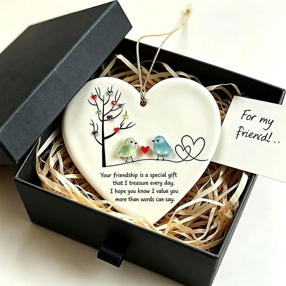 A heart-shaped ceramic friendship ornament displayed inside an open black gift box with shredded paper. The ornament is white with a printed tree, two small birds, and a quote, with a gold hanging loop and a small card reading 'For my friend!'.