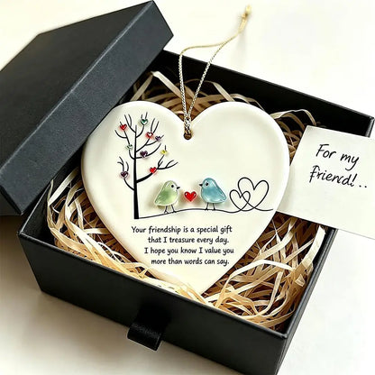 A heart-shaped ceramic friendship ornament displayed inside an open black gift box with shredded paper. The ornament is white with a printed tree, two small birds, and a quote, with a gold hanging loop and a small card reading 'For my friend!'.
