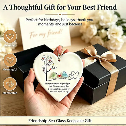 A hand holds a heart-shaped sea glass keepsake ornament featuring two birds and a tree, with a black gift box and ribbon in the background. Text overlays describe the thoughtful friendship gift.