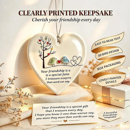 Heart-shaped ceramic keepsake plaque with a bird and tree illustration and a printed friendship message, displayed with a gift box, string lights, and decorative text on a warm beige background.
