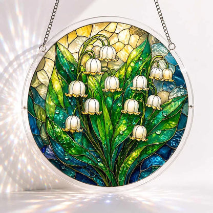 A round acrylic suncatcher showing white lily of the valley bells with green leaves and water droplets on teal glass