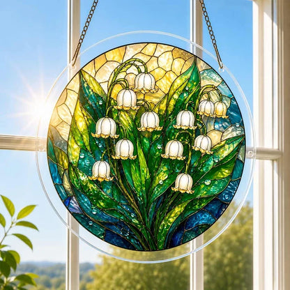 A round acrylic suncatcher with white lily of the valley bells and green leaves hanging from a chain in front of a bright white window