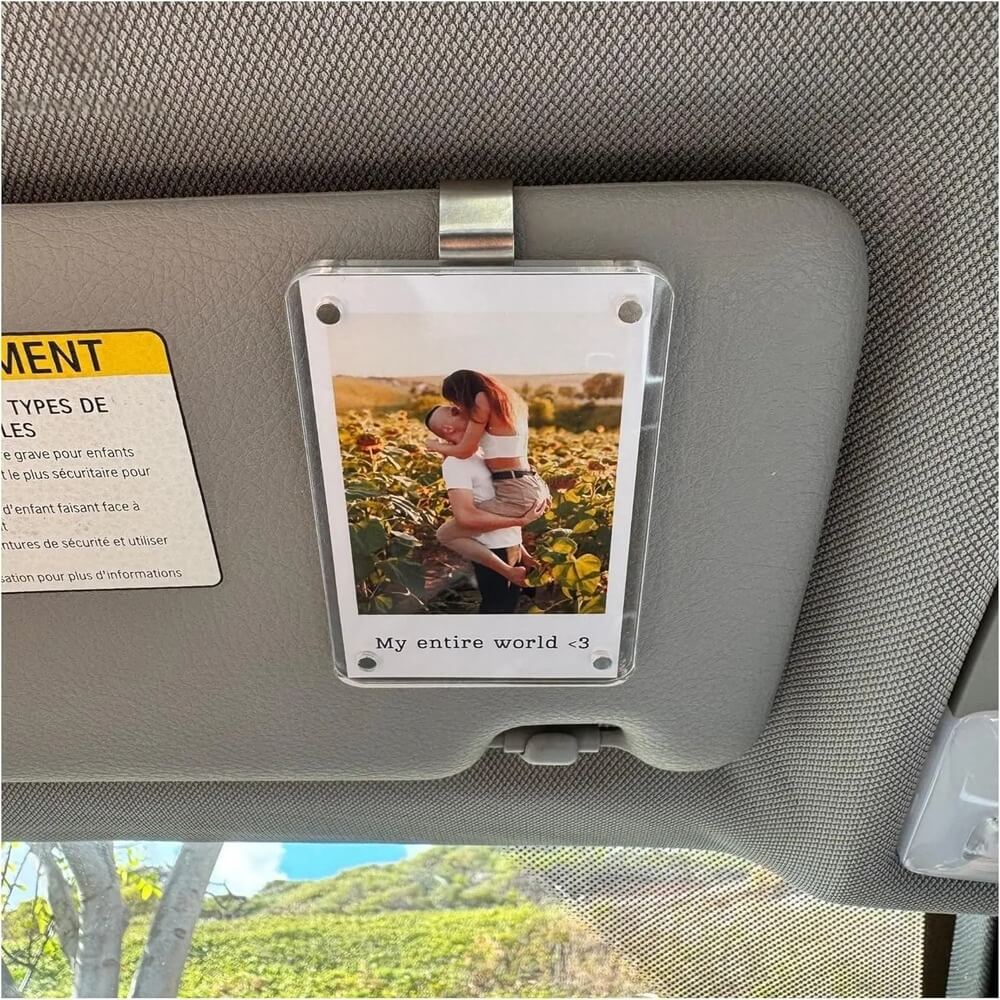 A clear acrylic car sun visor photo holder mounted on a gray visor inside a vehicle. The holder contains a printed photo of a woman carrying a child in a field, with the text 'My entire world <3' at the bottom.
