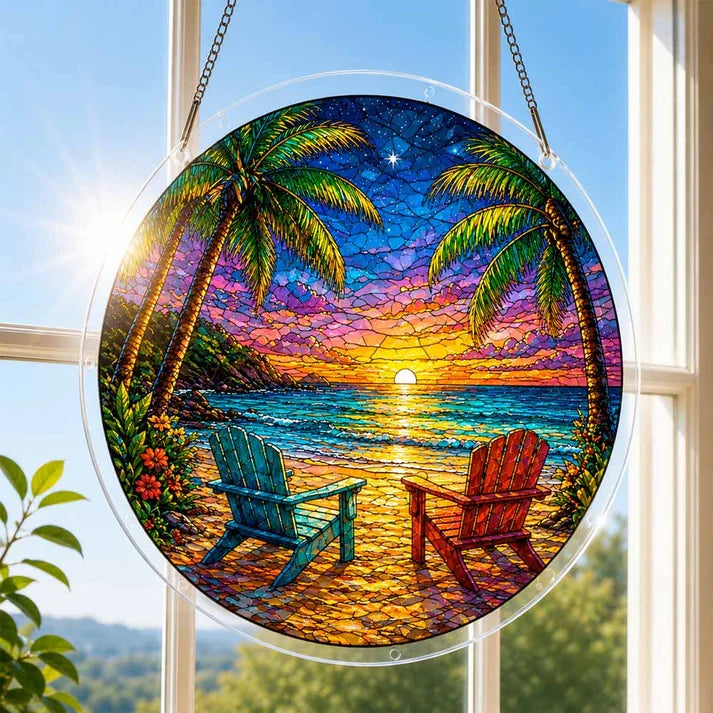 A round acrylic suncatcher with a tropical beach sunset two adirondack chairs and palm trees hanging from a chain in front of a bright white window