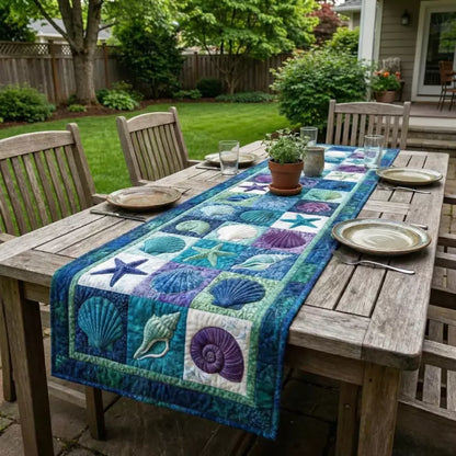 A quilted table runner with shells starfish and ocean squares on a wooden table on a patio surrounded by green lawn trees and a fence