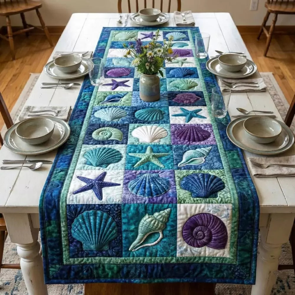 A quilted table runner with shells starfish and ocean patchwork squares on a white wooden table with bowls glasses cutlery and a vase of wildflowers