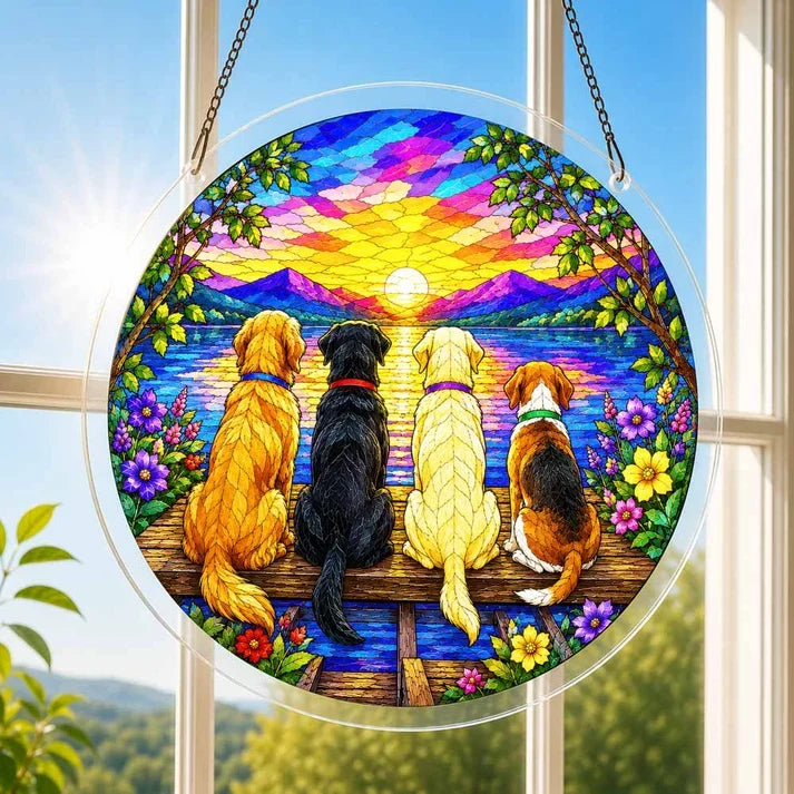 A round acrylic suncatcher with four dogs sitting on a wooden dock watching a sunset hanging from a chain in front of a bright white window