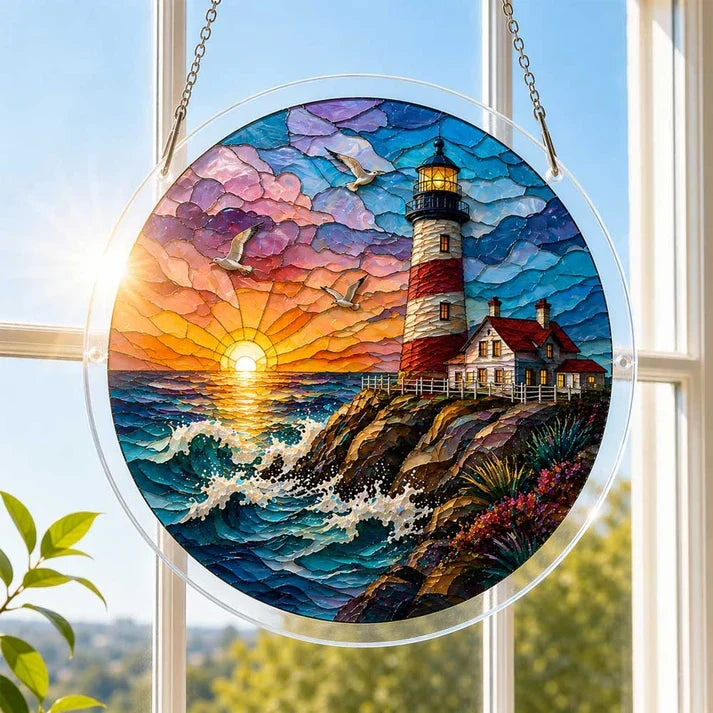 A round acrylic suncatcher with a red and white lighthouse at seaside sunset hanging from a chain in front of a bright white window