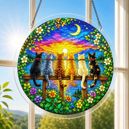 A round acrylic suncatcher with six cats sitting on a wooden fence watching a sunset hanging from a chain in front of a bright white window