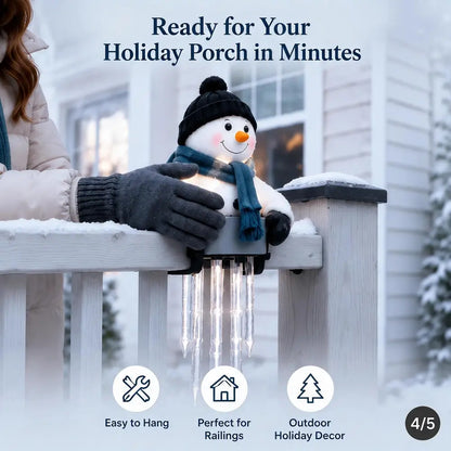 A snowman-shaped holiday porch light with a black hat, blue scarf, and glowing face is clipped onto a white railing in a snowy outdoor setting. A person's gloved hand is holding it in place.